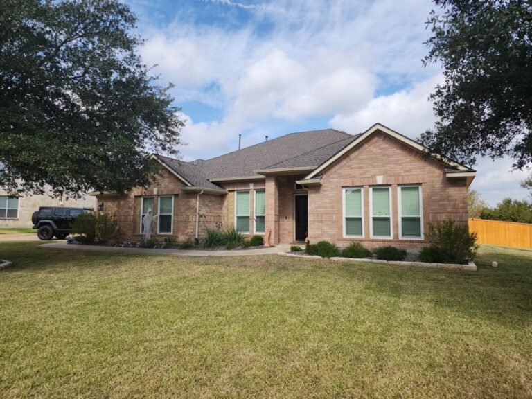 Front view of a single-story brick house with a spacious lawn and a black SUV parked on the driveway under a blue, partly cloudy sky.