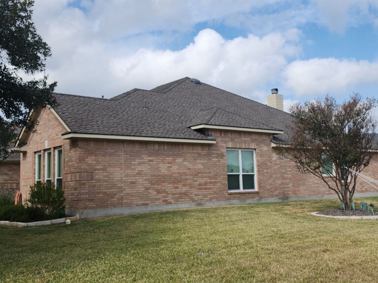 Brick suburban house with a brown shingled roof, green-framed windows, and a manicured front yard under a partly cloudy sky.