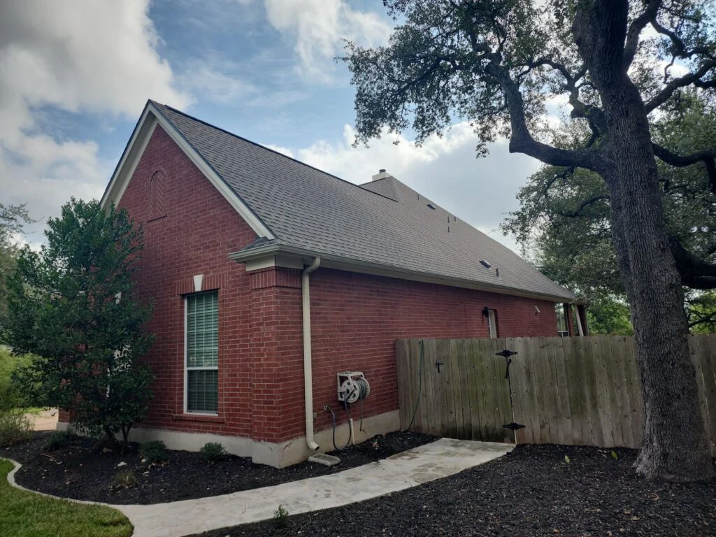 Red brick house with a steep gray roof, large tree to the right, and a curved concrete walkway in a backyard.
