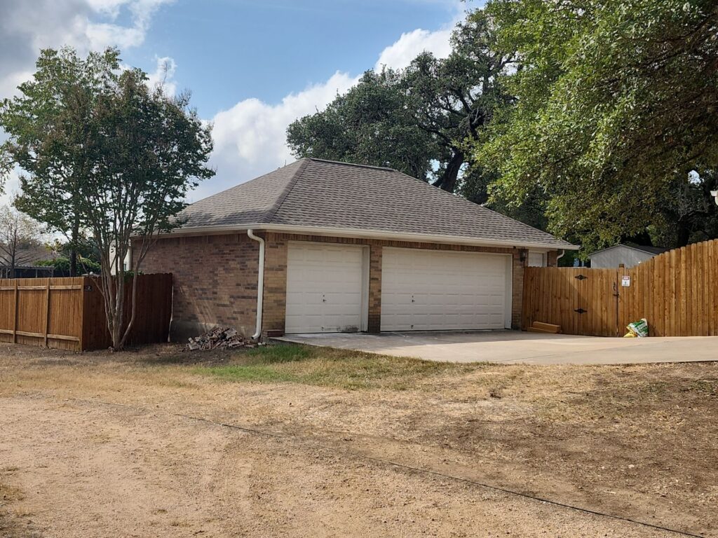 Two-car brick garage with brown shingled roof, surrounded by a wooden fence and mature trees under a blue sky.
