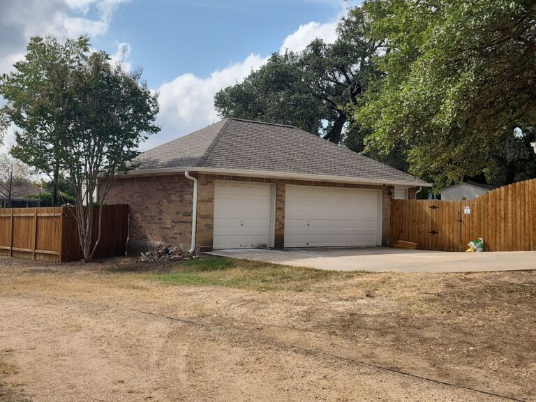 Two-car brick garage with brown shingled roof, surrounded by a wooden fence and mature trees under a blue sky.