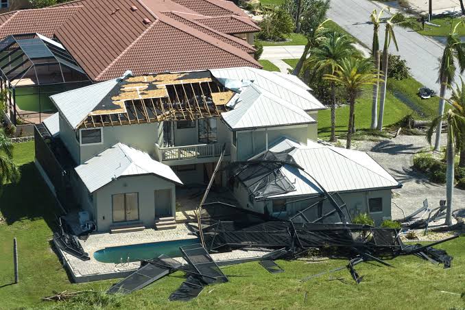 Two-story suburban house with major roof collapse and debris scattered across the yard, next to a small pool.