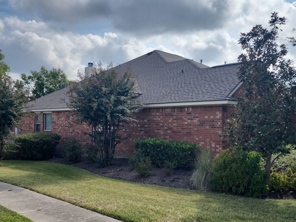 Brick suburban house with a gray, pitched roof, front yard landscaping, and a sidewalk in the foreground under a cloudy sky.