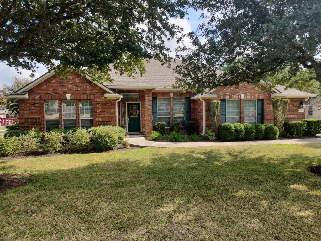Brick single-family home with a green front door, front yard shrubs, and overhanging tree branches.
