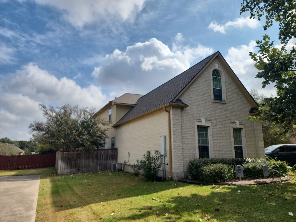 Brick house with a steep gable roof, arched window, and a front lawn under a blue sky with clouds; a car is parked on the right edge.