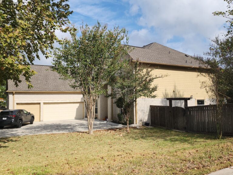 Suburban house with beige brick and 3-car? garage doors, front yard with trees, and a wooden fence on the right under a blue sky.