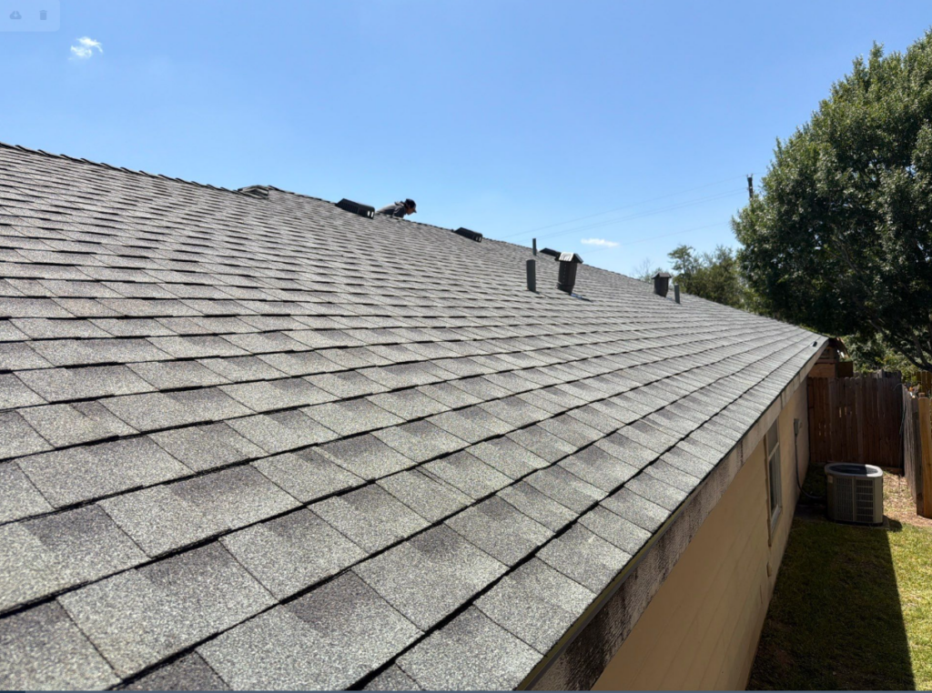 Sloped gray asphalt shingle roof with a person near the ridge inspecting vents; side yard with a fence and air conditioner under a clear blue sky.