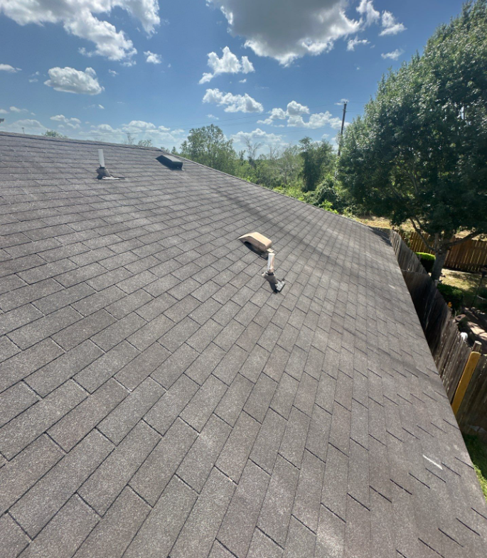 Gray shingle roof viewed from above with two vent caps; trees and a wooden fence visible in the background under a blue sky.