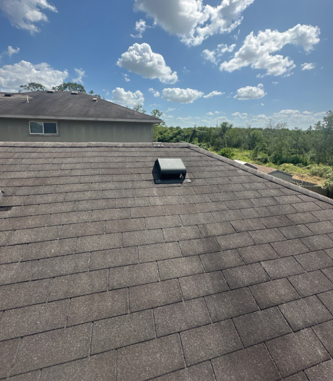 before-wide-view-existing-roof-weathered-shingles-caleb-drive - Driftwood Builders Roofing Residential rooftop with dark asphalt shingles, a small roof vent near the center, and a clear blue sky with fluffy white clouds; distant treeline visible in the background.