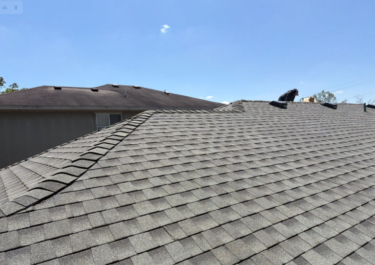 after-completed-gaf-timberline-hdz-hip-ridge-cap-caleb-drive - Driftwood Builders Roofing Gray shingled roof with workers on the ridge under a clear blue sky. Looks like roof repair or installation in progress far from ground level.