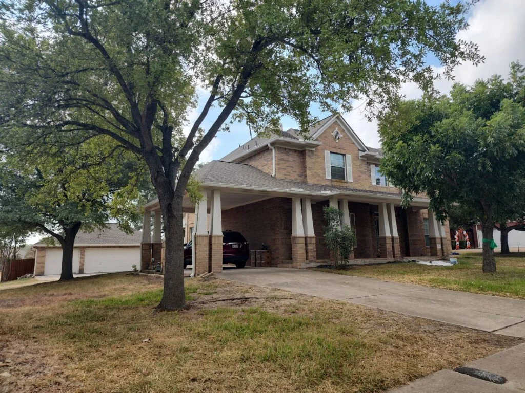Two-story brick house with a covered front porch, white columns, and a large tree in the yard shading the driveway with a parked SUV underneath the carport edge.