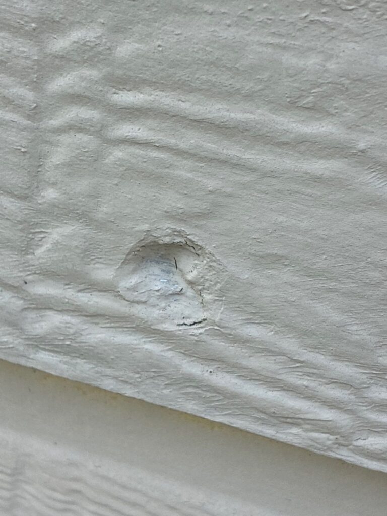Close-up of a hail impact chip in painted fibre cement siding, exposing the material beneath the finish