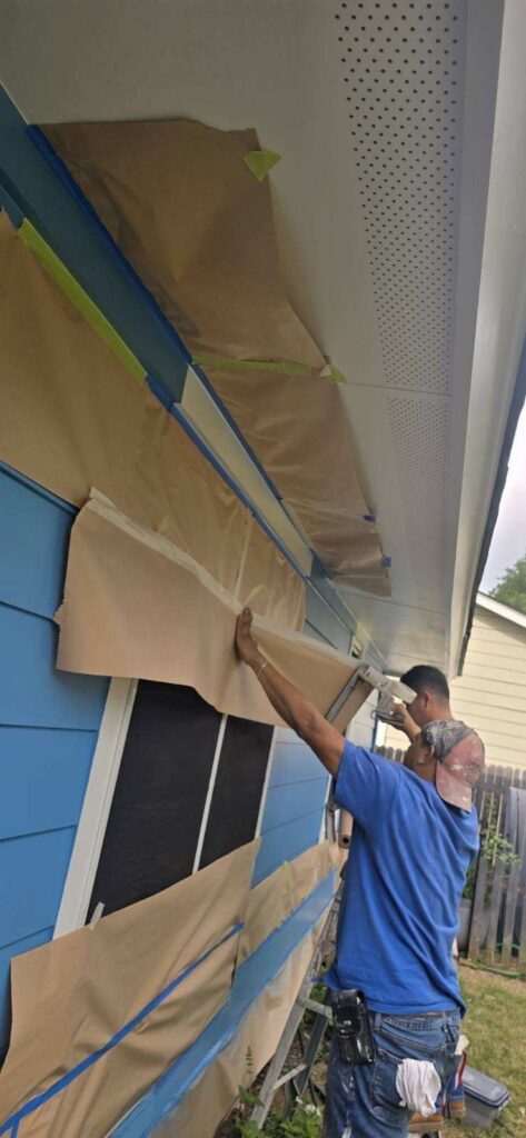 A worker on a ladder masks blue house siding with brown paper and painter's tape for painting or prep.