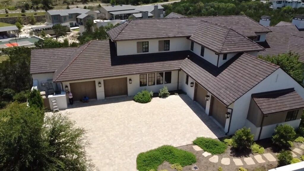 A large white house with a brown tile roof and multiple garage doors, viewed from above, with a spacious tiled courtyard and surrounding landscaping.