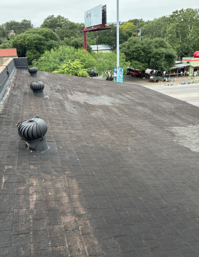 Aerial view of a flat, dark shingle roof with three large round exhaust vents, beyond which a street and billboards are visible among green trees. - Driftwood Builders Roofing Aerial view of a flat, dark shingle roof with three large round exhaust vents, beyond which a street and billboards are visible among green trees.