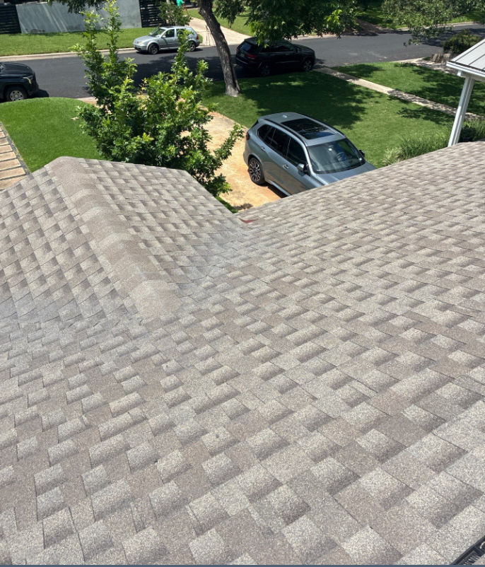Aerial view from a roof looking down onto a suburban street: gray shingled roof in the foreground, a silver SUV parked on a gravel driveway, with green lawns and trees nearby.