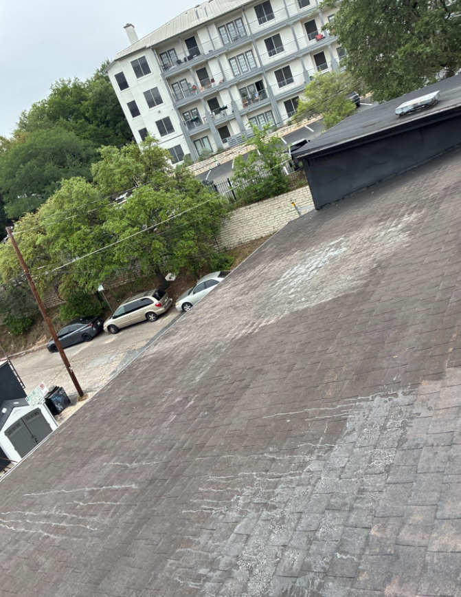 Tilted rooftop with weathered shingles, showing a street with parked cars and a white multi-story apartment building in the background. - Driftwood Builders Roofing Tilted rooftop with weathered shingles, showing a street with parked cars and a white multi-story apartment building in the background.