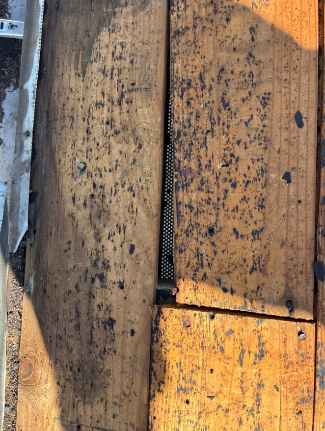 Close-up of weathered wooden boards with dark stains and a narrow gap revealing perforated metal behind.