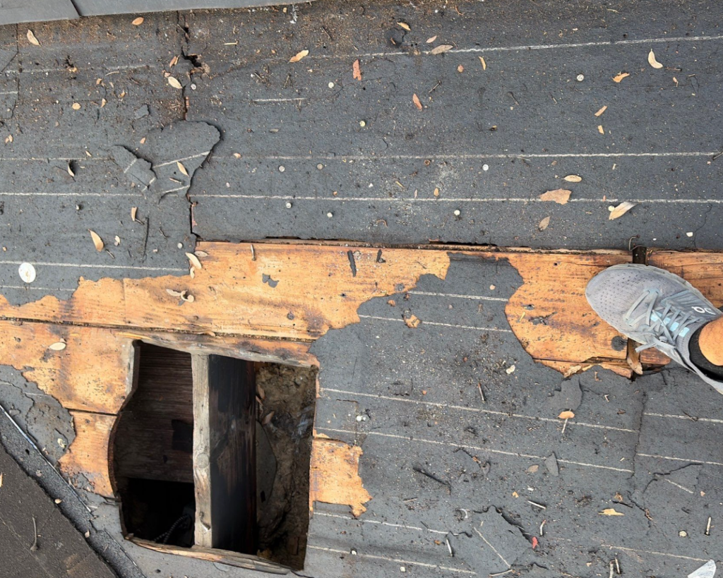 Open hole in a damaged roof with exposed wood and torn underlayment; a sneaker rests at the edge nearby.