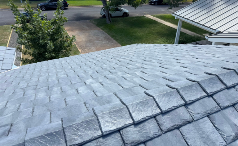 Close-up aerial view of gray slate roof shingles, with a residential street and trees visible in the background.
