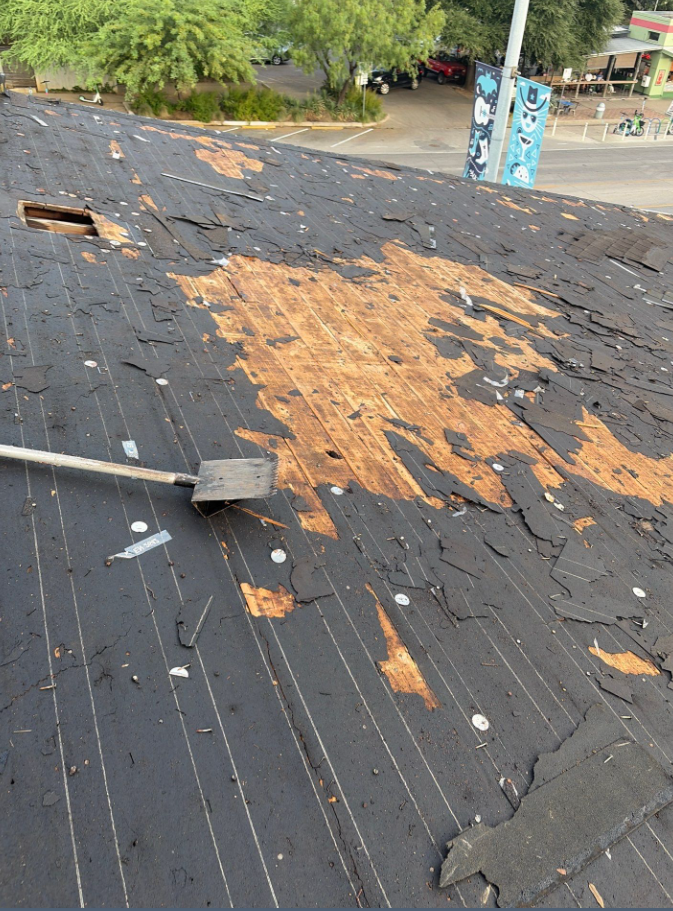 Rooftop with peeling asphalt shingles exposing wooden underlayment after damage; a long-handled tool rests on the sloped roof. - Driftwood Builders Roofing Rooftop with peeling asphalt shingles exposing wooden underlayment after damage; a long-handled tool rests on the sloped roof.