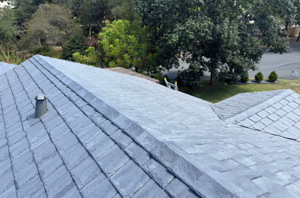 Gray slate roof with a vent pipe in the foreground and a ladder resting against the peak; suburban street and trees in the background