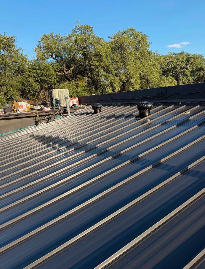 Metal standing-seam roof with two circular vents and scattered construction materials along the edge, backed by trees and a clear blue sky. - Driftwood Builders Roofing Metal standing-seam roof with two circular vents and scattered construction materials along the edge, backed by trees and a clear blue sky.