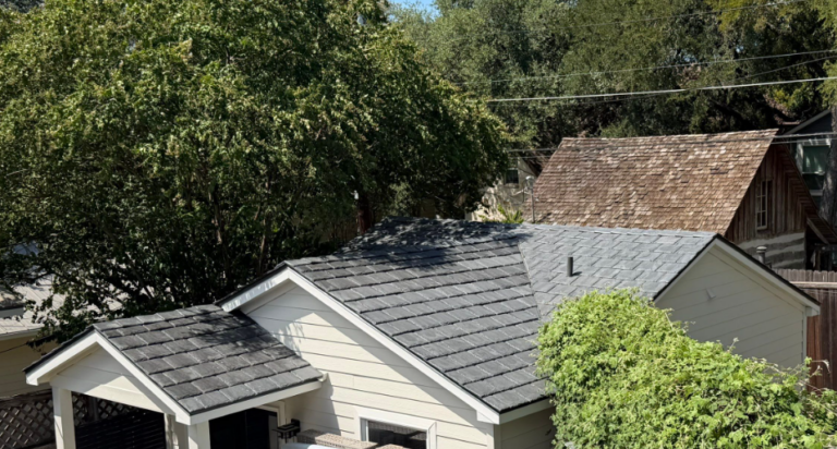 Row of suburban houses with gray shingled roofs and green trees in the background, sunny daylight.