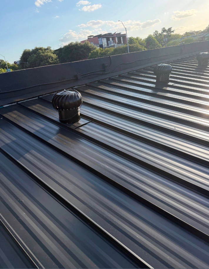 Metal standing-seam roof with two black round vent caps; blue sky, trees, and a building visible in the background. - Driftwood Builders Roofing Metal standing-seam roof with two black round vent caps; blue sky, trees, and a building visible in the background.