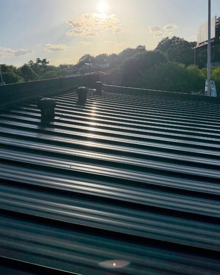 Sun glare over a corrugated metal rooftop with vents, trees in the background. - Driftwood Builders Roofing Sun glare over a corrugated metal rooftop with vents, trees in the background.