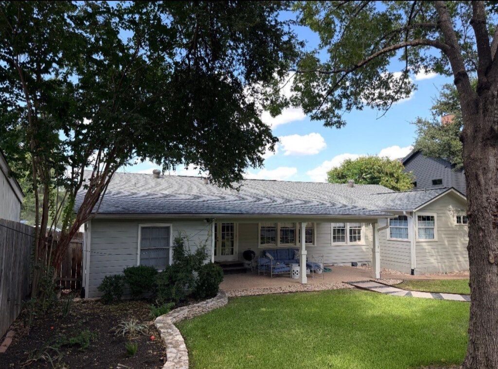 Backyard of a single-story home with a covered patio, blue seating, and a green lawn under a tree-lined sky.
