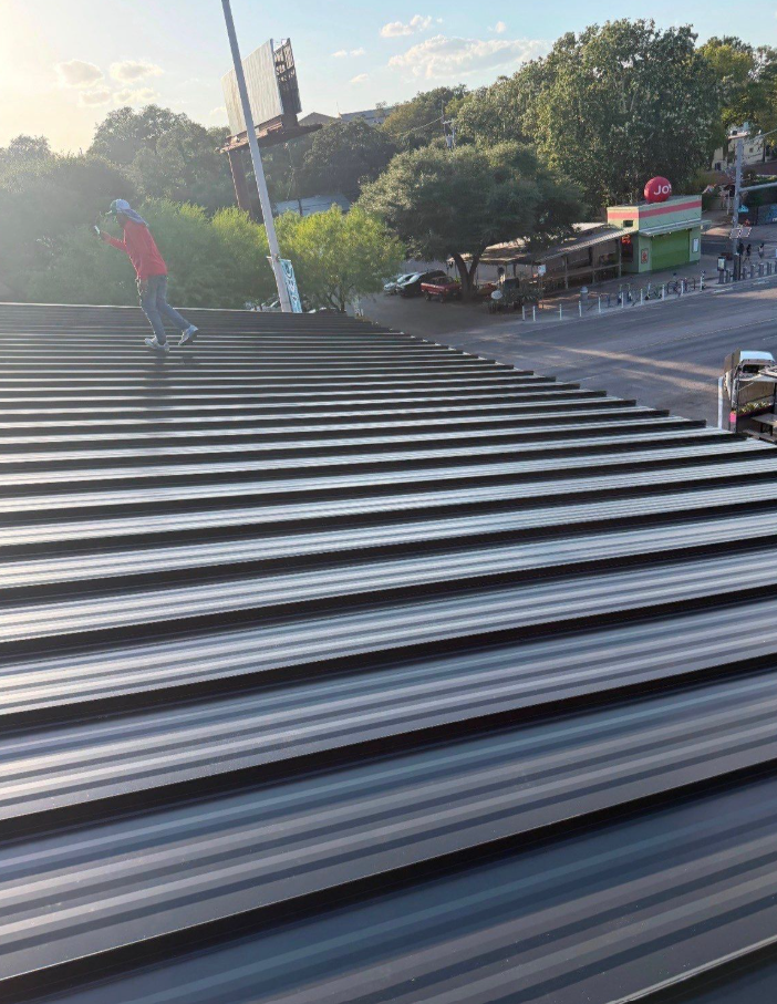 Person wearing a pink shirt and jeans walks along a rooftop with corrugated metal panels, city streets and trees in the background.