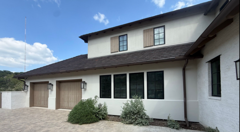 Two lighted garage doors and a white house exterior with three black-framed windows, shrubs, and a paved driveway on a sunny day.