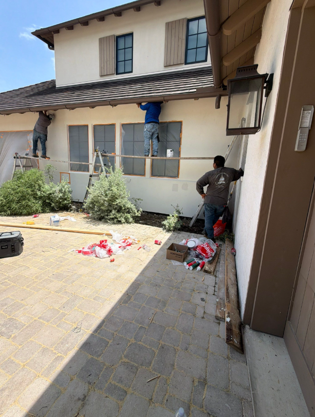 Three construction workers on a scaffold and ground outside a beige two-story house, doing exterior work under the roof eaves.