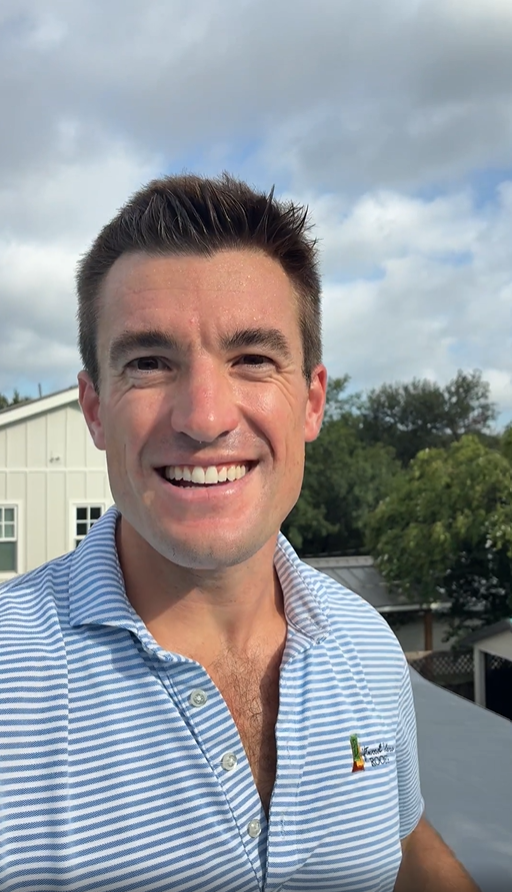 Smiling man in a light blue striped polo on a rooftop deck with trees and houses in the background, on a sunny day.