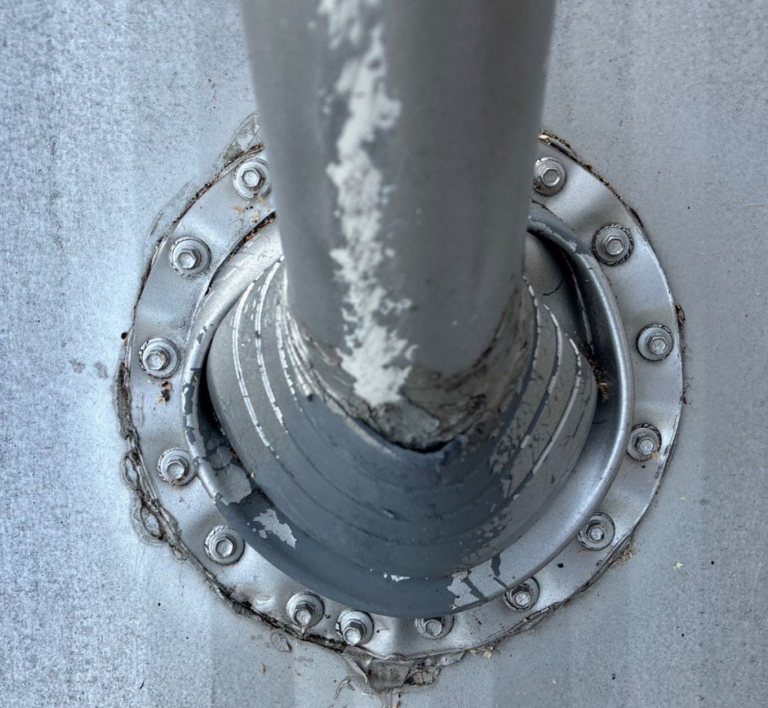 Close-up of a weathered metal pipe passing through a circular bolt-flange on a gray surface, with peeling paint and rust around the joint.