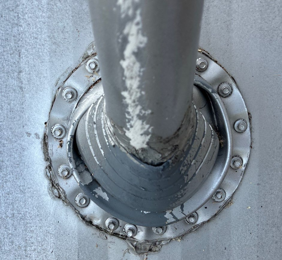Close-up of a weathered metal pipe passing through a circular bolt-flange on a gray surface, with peeling paint and rust around the joint.
