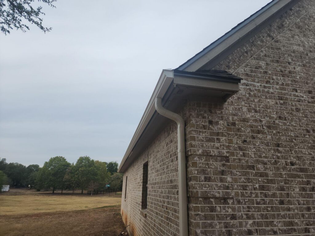 Brick house exterior with white gutter and downspout along the side, sloped roof, and a grassy, tree-lined yard in the distance under a cloudy sky.