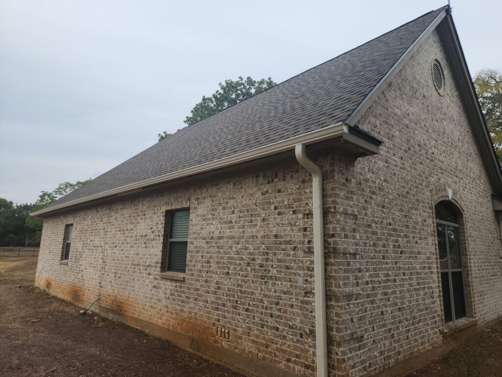 Brick house with steep gray-shingled roof, white gutter running down the wall, and brick exterior siding visible side angle.