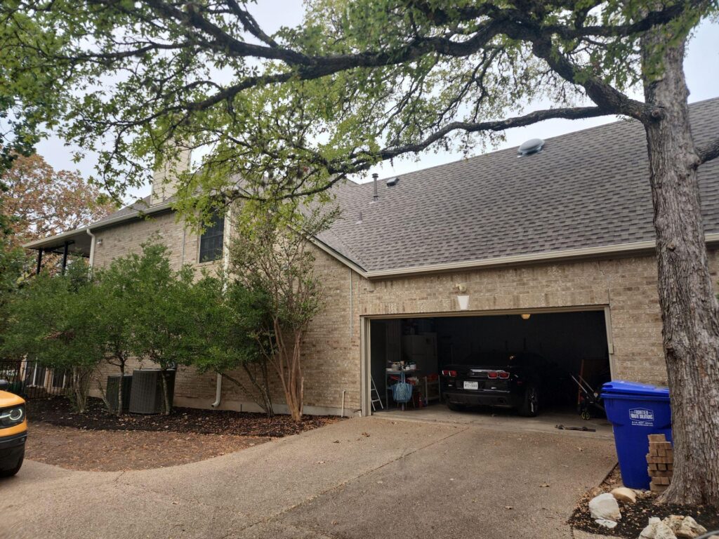 Brick house with an open garage showing a car inside; trees and shrubs frame the driveway, with a blue recycling bin nearby.