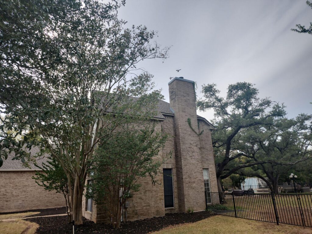 Brick house with a tall chimney surrounded by trees and a black metal fence in a suburban yard; cloudy sky above, birds in flight near the chimney.