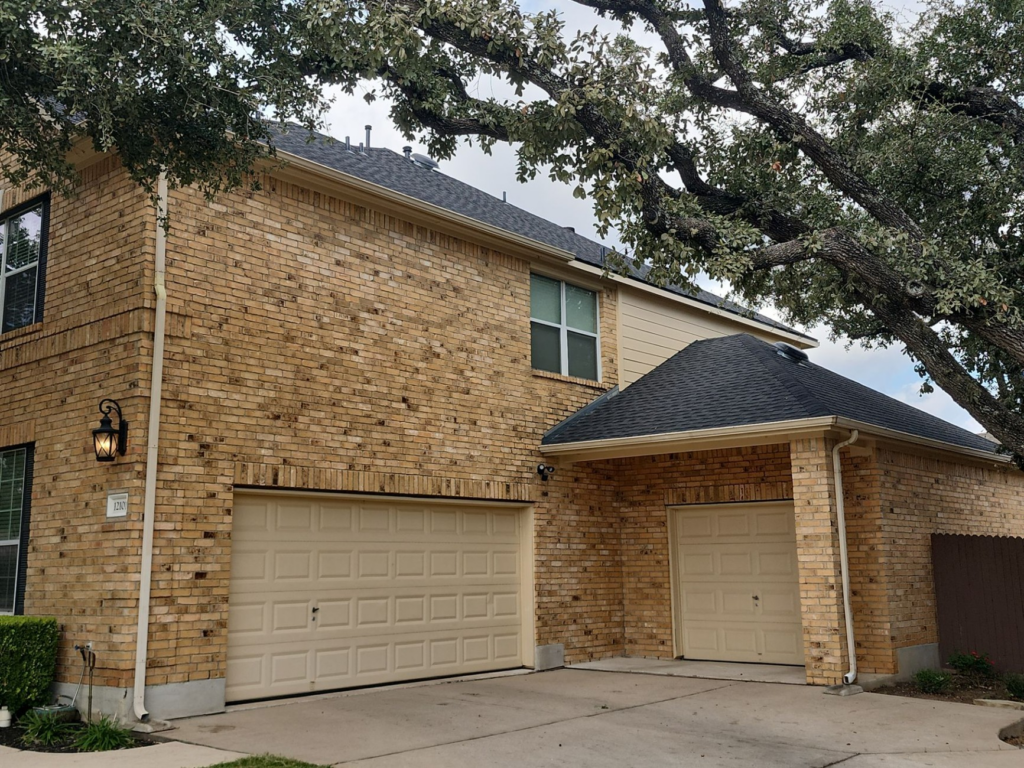 Two-story brick townhouse with twin beige garage doors and a large tree arching over the roof.