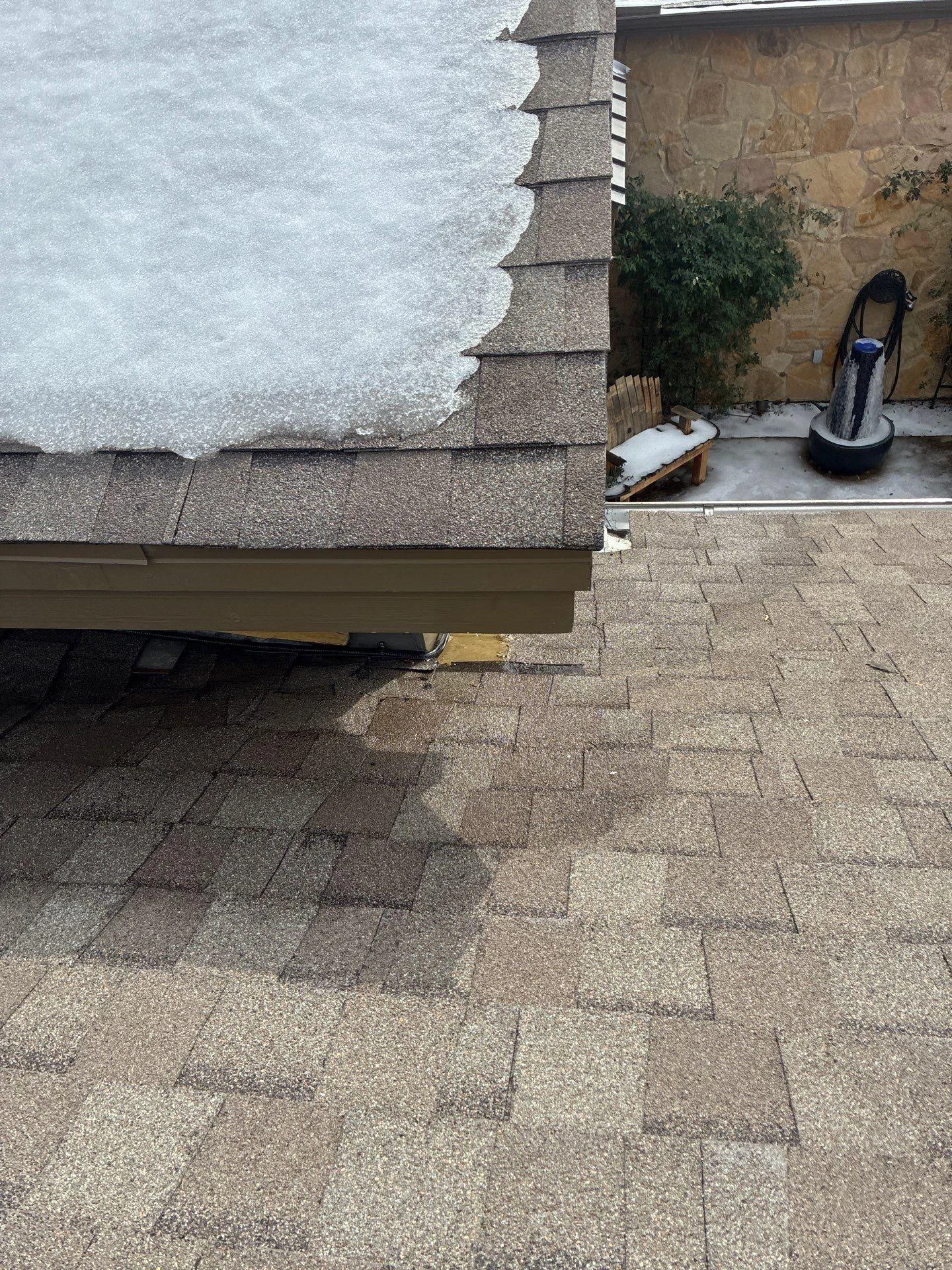 Overhead view of a brown shingle roof with snow along the left edge and a stone-walled patio area to the right.