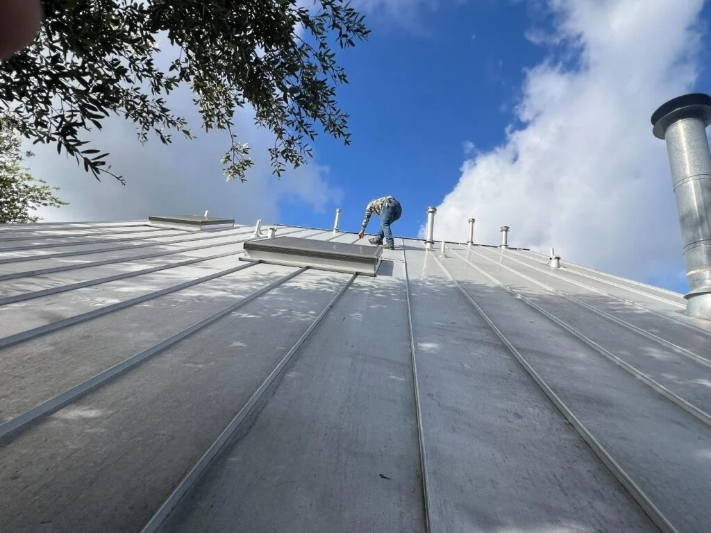Worker on a sloped metal roof installing or inspecting vents under a blue sky with clouds; tree branches visible at left.