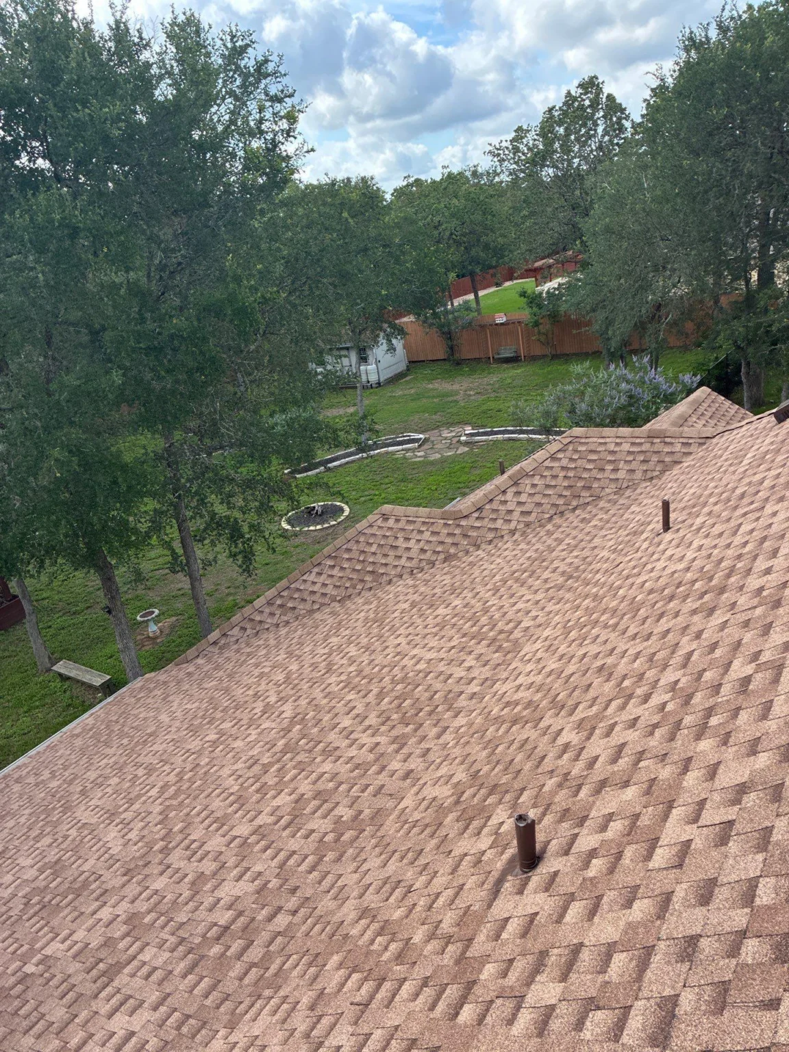 Aerial view from a brown shingled roof of a backyard with a circular garden bed, birdbath, trees, and a wooden fence.