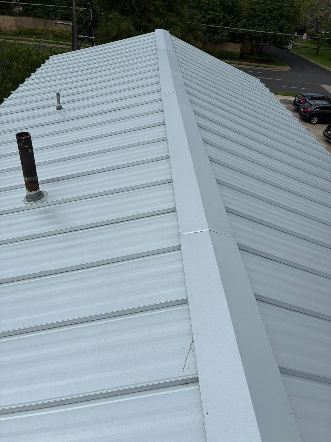 Overhead view of a light gray metal standing-seam roof with a central ridge and two roof vents/pipes nearby a street and parking area.