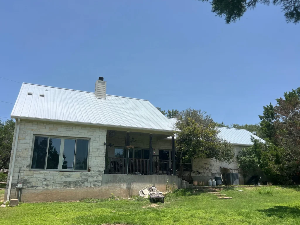 Front view of a single-story stone house with a white metal roof and a covered front porch on a sunny day, with a grassy yard in the foreground.