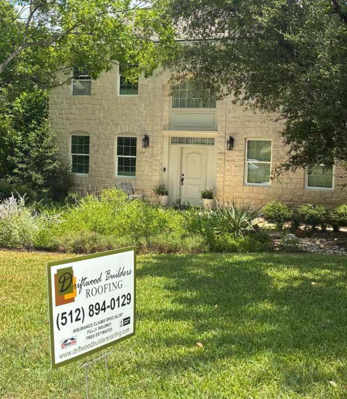 Front of a beige stone house with a landscaped yard and a roofing sign in the lawn.