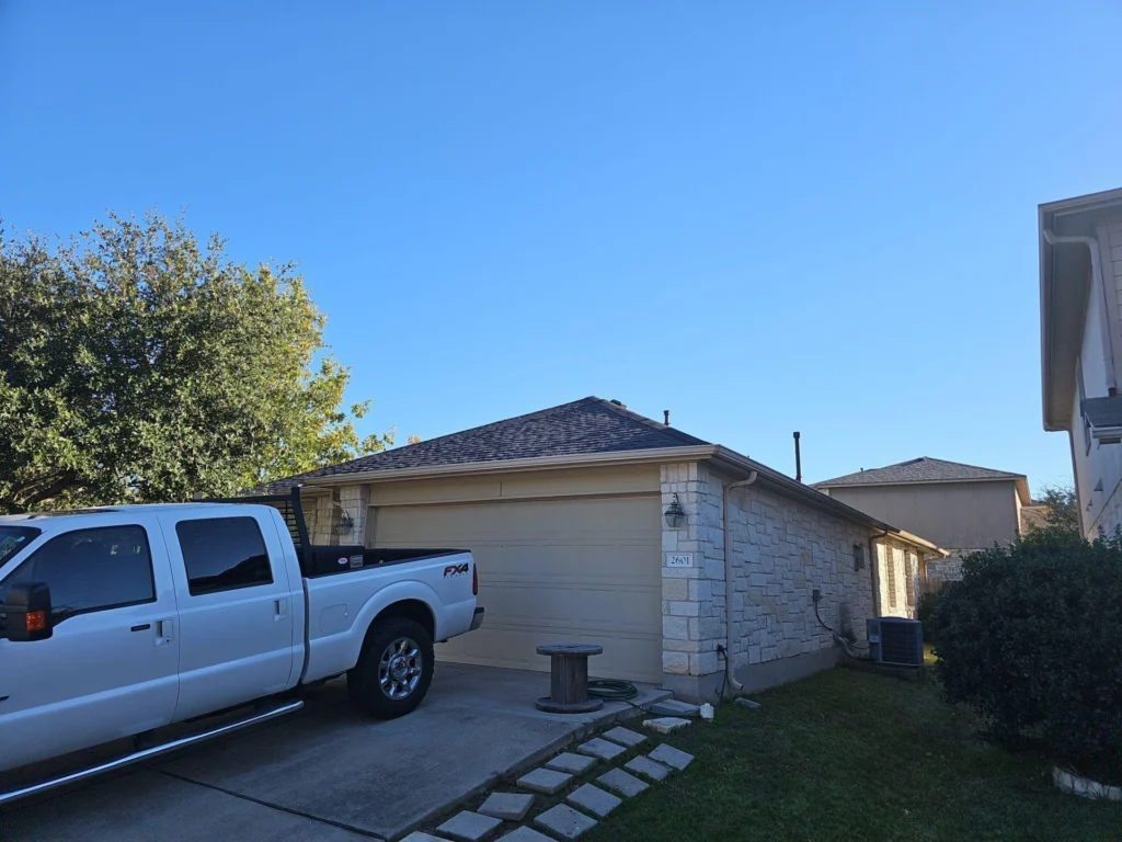 White crew-cab pickup truck parked in a driveway in front of a beige stone-faced garage at a suburban home under a clear blue sky.