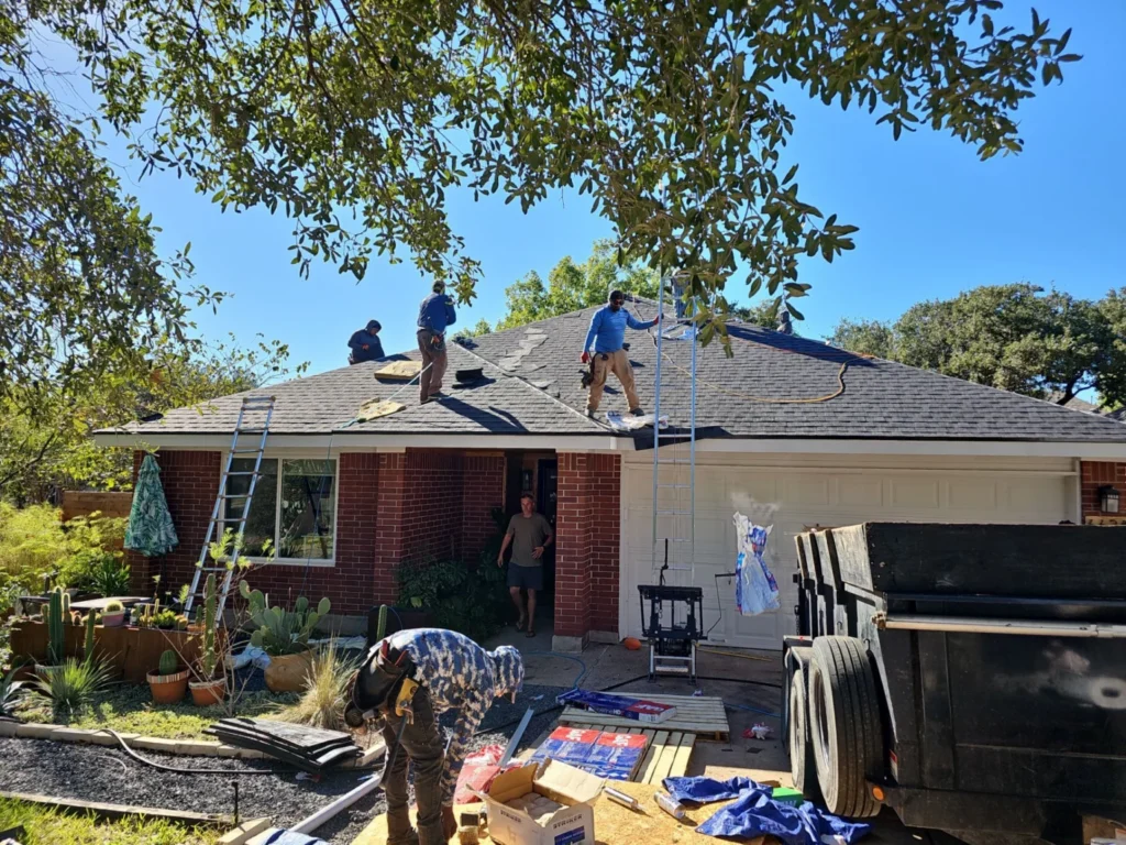Crew installing new shingles on a single-story brick house roof, workers on the roof with ladders and supplies nearby on a sunny day.